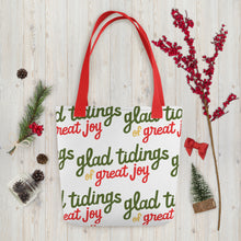 Load image into Gallery viewer, A white tote bag with red handles laying on a table with Christmas items around it. The tote bag features the words &quot;glad tidings of great joy&quot; in green, yellow and red. 