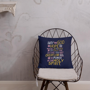 A pillow on a chair with a coffee mug on a table next to it. The purple pillow features hand drawn lettering of the Bible verse "May the God of hope fill you with all joy and peace as you trust him, so that you may overflow with hope by the power of the holy spirit." The lettering in white, pink and yellow. 