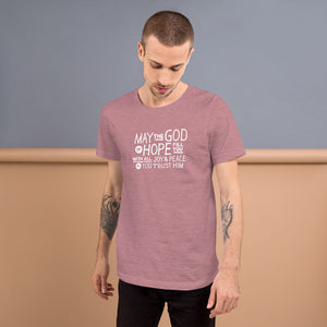 A man wearing an orchid pink color short sleeved t-shirt. The t-shirt features hand drawn lettering in white with the words "May the God of hope fill you with all joy and peace as you trust him."