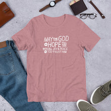 Load image into Gallery viewer, A short sleeved T-shirt laying flat with objects around it. The tee is a orchid pink color and features hand drawn lettering in white with the words &quot;May the God of hope fill you with all joy and peace as you trust him.&quot;