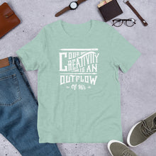Load image into Gallery viewer, A short sleeved T-shirt laying flat with objects around it. The tee is a Heather Prism Dusty Blue color and features hand drawn lettering in white with the words &quot;Our creativity is an outflow of His.&quot;