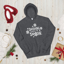 Load image into Gallery viewer, A hoodie laying on the ground with Christmas decorations and red slippers around it. The dark grey hoodie features the words Dashing through the snow in the middle in white. There are snowflake illustrations surrounding the words also in white. 