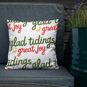 The white pillow is leaning on a sofa with a plant off to the side. The white pillow has a repeat pattern with the phrase "glad tidings of great joy" in the colors green, red and yellow. 