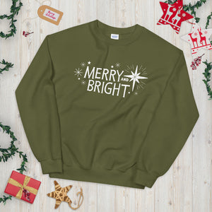 A hunter green sweatshirt laying on a table with Christmas objects around it. The sweatshirt features the words Merry and Bright with illustrated Christmas stars around it. The words and illustrations are in white on the hunger green fabric. 
