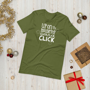 A olive green T-shirt laying on the ground with Christmas items surrounding it. The T-shirt features the words  "Up on the housetop, click, click, click" in white. There are three stars around the words in white. 