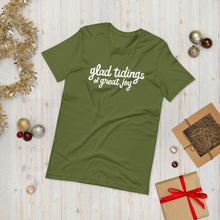 Load image into Gallery viewer, An olive green T-shirt laying on the ground with Christmas items surrounding it. The T-shirt features the words  &quot;glad tidings of great joy&quot; in white. 