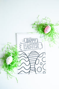 A coloring sheet on a white tabletop with fake green Easter grass and Easter eggs around the sheet. The coloring sheet features the words “Happy Easter” with illustrated Easter eggs.