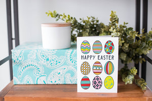 A greeting card is on a table top with a present in blue wrapping paper in the background. On top of the present is a candle and some greenery from a plant too. The card features illustrated Easter eggs in bright fun colors with the words “Happy Easter” in the middle of the eggs.