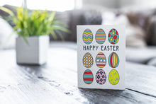 Load image into Gallery viewer, A greeting card featured on a black, wood coffee table. There’s a white planter in the background with a green plant. There’s also a gray sofa in the background with a white pillow. The card features illustrated Easter eggs in bright fun colors with the words “Happy Easter” in the middle of the eggs. 