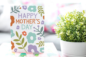 A photo of a card featured on a tabletop next to a white planter filled with a green plant. ​​The card features the words “Happy Mother’s Day” with illustrated flowers around the words. 