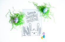 Load image into Gallery viewer, A coloring sheet on a white tabletop. There’s fake Easter grass around the color page. The coloring page features an illustrated Easter bunny with some leaves around the bunny. The words “have a hopping Easter” are above the bunny. 