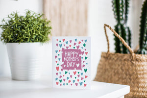 A greeting card is featured on a white tabletop with a white planter in the background with a green plant. There’s a woven basket in the background with a cactus inside. The card features the words “Happy Mother’s Day.”
