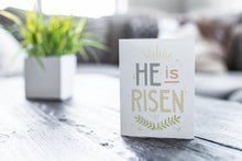 Load image into Gallery viewer, A greeting card featured on a black, wood coffee table. There’s a white planter in the background with a green plant. There’s also a gray sofa in the background with a white pillow. The card features the words “He is Risen” with palm leaves at the bottom of the design. 