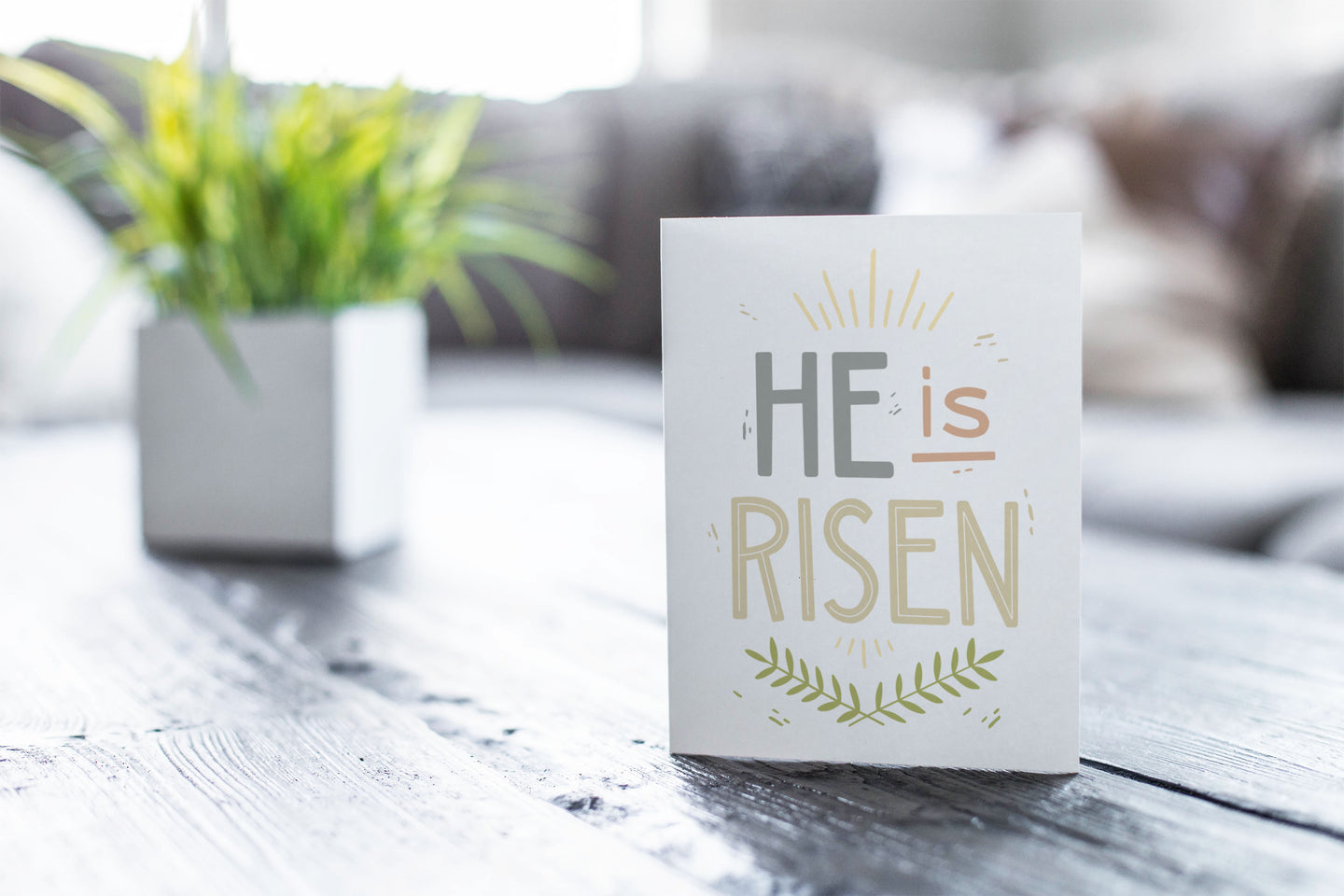 A greeting card featured on a black, wood coffee table. There’s a white planter in the background with a green plant. There’s also a gray sofa in the background with a white pillow. The card features the words “He is Risen” with palm leaves at the bottom of the design. 