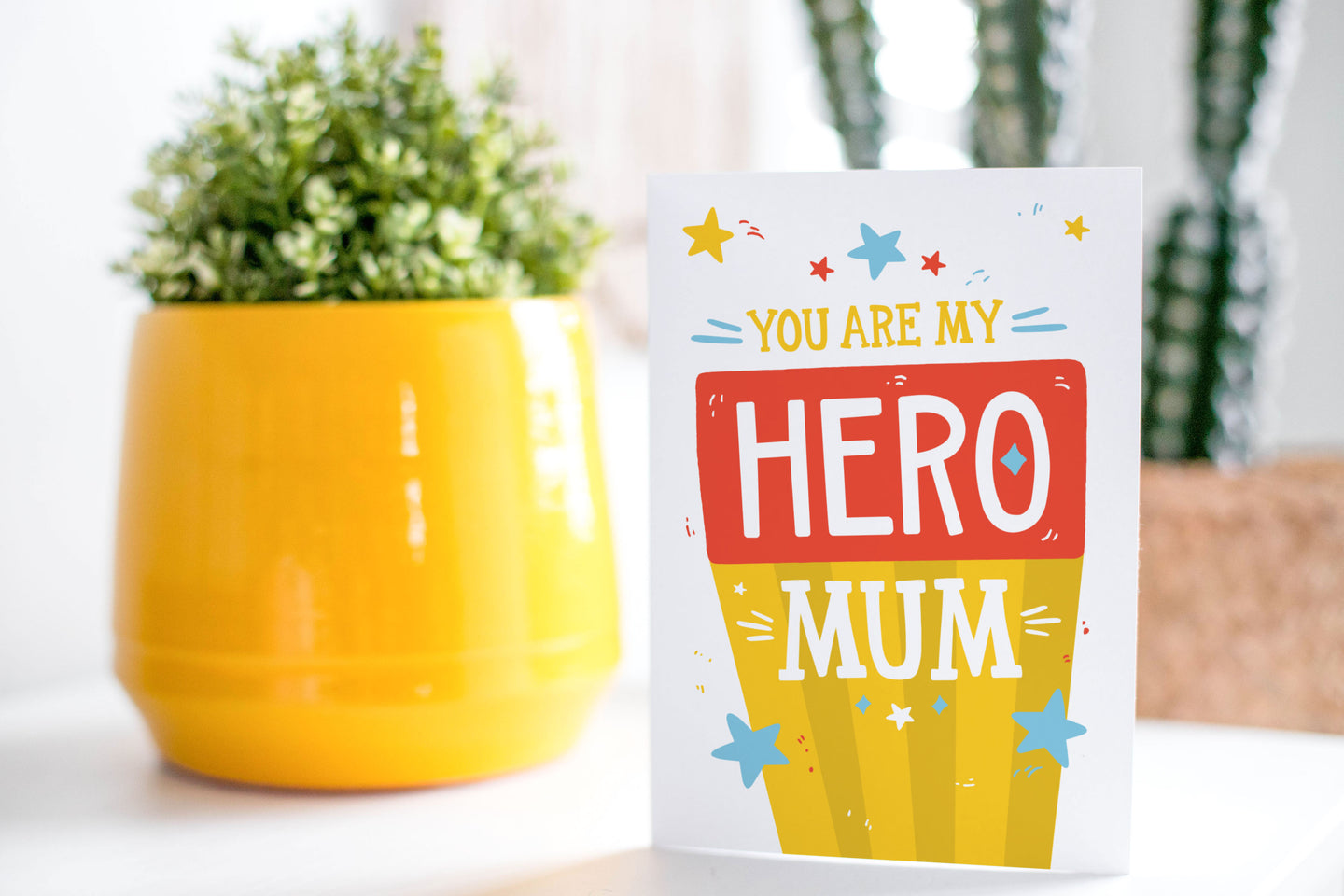 A greeting card is on a table top with a yellow plant pot and a green plant inside. The card features illustrated lettering reading “You are my hero mum” with stars around it. There’s a background behind the word “mum” featuring yellow stripes and the word “hero” has a red background. 