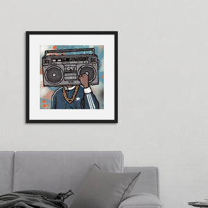 A black frame on a wall above a grey sofa. In the frame is an illustrated image of a boombox with a person wearing a gold chain and blue jacket. 