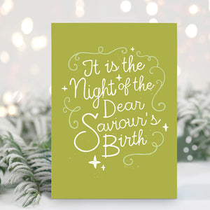 A Christmas card standing up with with pine leaves in the background with a touch of snow. The background of the card is a lime green with the word "it is the night of the dear saviour's birth" in script white lettering.