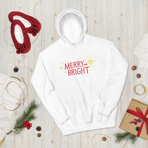 A hoodie laying on  the ground with Christmas decorations and red slippers around it. The white hoodie features the words Merry and Bright in the middle in red. Around the words are yellow illsutrated stars. 