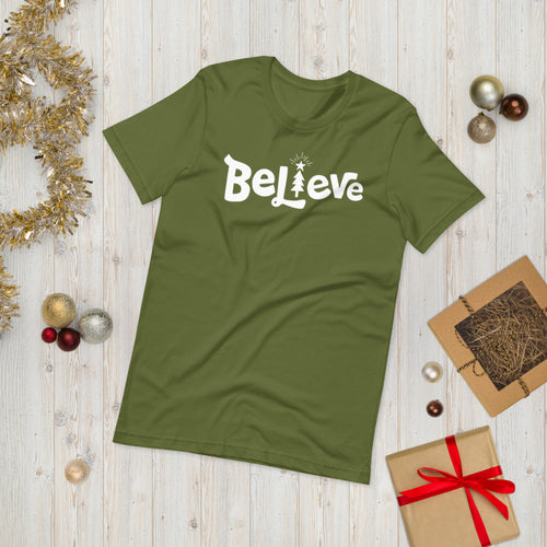 A olive green T-shirt laying on the ground with Christmas items surrounding it. The T-shirt features the word Believe in the middle in white lettering with the 