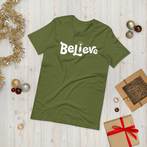 A olive green T-shirt laying on the ground with Christmas items surrounding it. The T-shirt features the word Believe in the middle in white lettering with the "I" of the word featured as a illustrated Christmas tree. 