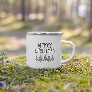 A white enamel mug with a silver enamel rim sitting in the grass. The illustrated design says "Merry Christmas" with illustrated trees below. The design is in black. 