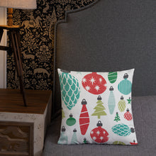 Load image into Gallery viewer, A pillow leaning on a grey headboard with a table and lamp off to the side. The white pillow features Christmas illustrations with vintage ornaments. The ornaments are featured in blue, red and green. 