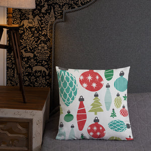 A pillow leaning on a grey headboard with a table and lamp off to the side. The white pillow features Christmas illustrations with vintage ornaments. The ornaments are featured in blue, red and green. 