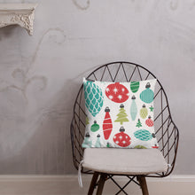 Load image into Gallery viewer, The pillow is leaning on a metal chair with a cushion. The white pillow features Christmas ornament illustrations in the colors red, green and blue. 
