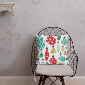 The pillow is leaning on a metal chair with a cushion. The white pillow features Christmas ornament illustrations in the colors red, green and blue. 