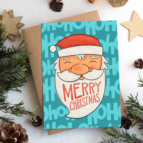 A photo of a Christmas card on top of a brown paper wrapped gift with Christmas decor around it. The card has a blue background with the words 'ho ho ho' in a lighter shade of blue. On top of the background is an illustrated Santa Claus with the words 