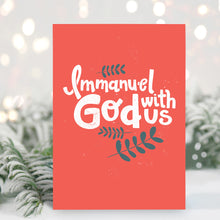 Load image into Gallery viewer, A Christmas card standing up with with pine leaves in the background with a touch of snow. The card has a light red background with the words &quot;Immanuel God with Us&quot; in white with a couple of plant leaves in navy around the words.