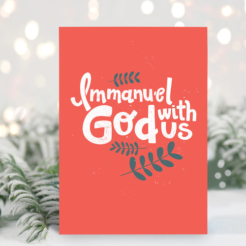 A Christmas card standing up with with pine leaves in the background with a touch of snow. The card has a light red background with the words 