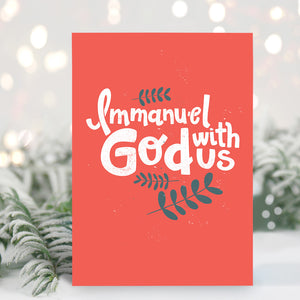 A Christmas card standing up with with pine leaves in the background with a touch of snow. The card has a light red background with the words "Immanuel God with Us" in white with a couple of plant leaves in navy around the words.