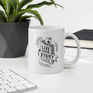 A mug featured on a desk with a plant and a keyboard. The mug is white with the artwork in black. The design features the words "Live Your Story" with the words inside an illustrated book. 