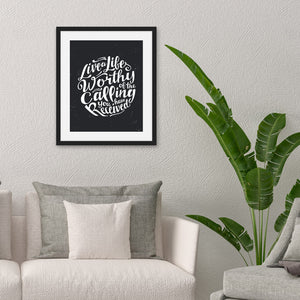 Lettering artwork is featured in a black frame above a sofa. The artwork is on a black background with some white texture to give a vintage look. The text is in white and reads “Live a life worthy of the calling you have received.” 