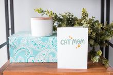 Load image into Gallery viewer, A greeting card is on a table top with a present in blue wrapping paper in the background. On top of the present is a candle and some greenery from a plant too. The card features the words “Cat mom.”
