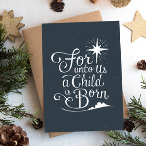 A photo of a Christmas card on top of a brown paper wrapped gift with Christmas decor around it. The Christmas card has a navy color as the background with white words reading "for unto us a child is born." Above the words is an illustrated Bethlehem star and below the words is a small illustration of the city of Bethlehem.