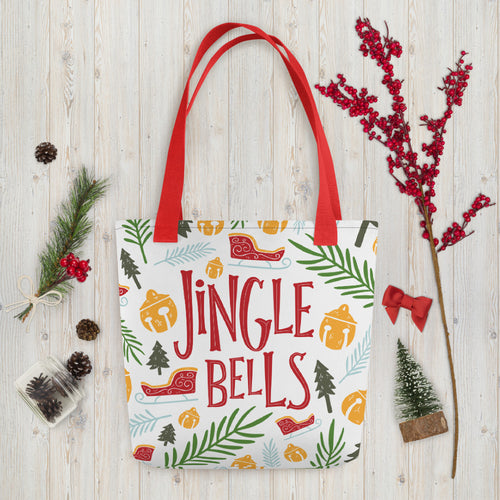 A white tote bag with red handles laying on a table with Christmas items around it. The tote bag features Christmas illustrations with the words Jingle Bells in the middle in red. The illustrations featured are ornaments, sleighs, pine trees and leaves. The illustrations are in the colors light and dark green, light blue, yellow and red. 
