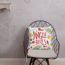 Load image into Gallery viewer, The pillow is leaning on a metal chair with a cushion. The white pillow features Christmas illustrations with the words Jingle Bells in the middle in red. The illustrations featured are ornaments, sleighs, pine trees and leaves. The illustrations are in the colors light and dark green, light blue, yellow and red. 