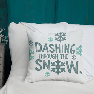 A white pillow with illustrations leading on white bedding with a side table off to the side. The white pillow has the words "Dashing through the snow" in light and dark blue in a repeating pattern. Around the words are illustrated snowflakes. 