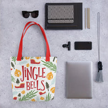Load image into Gallery viewer, A tote bag lying on a surface with a laptop and office items next to it. The Christmas tote bag features the words Jingle Bells in red with an illustrated Christmas pattern around the words. The illustrations are a sleigh, leaves, pine trees, and ornaments in the colors yellow, light blue, red, and light and dark green.