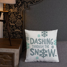 Load image into Gallery viewer, A pillow leaning on a grey headboard with a table and lamp off to the side. The white pillow is white with the words &quot;Dashing through the snow&quot; in light and dark blue with snowflakes. The words and snowflakes create a pattern on the pillow. 