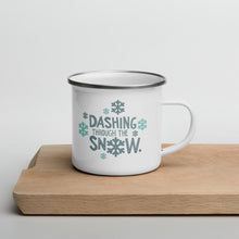 Load image into Gallery viewer, A white enamel mug sitting on top of a wood cutting board on a white background. The mug is white with the very top silver enamel. The design features the words &quot;Dashing through the snow&quot; in dark and light blue. There are snowflakes around the words. 
