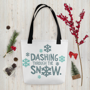 A white tote bag with black handles laying on a table with Christmas items around it. The tote bag features the words "Dashing through the snow" in light and dark blue with snowflakes around the words. 