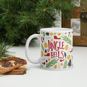 A white mug with a pine tree in the background. The mug feaures the words "Jingle Bells" with Christmas illustrations around the words. The illustrations include leaves, pine trees, ornaments and sleighs. The words are in red and the illustrations are in yellow, light blue, light and dark green. 