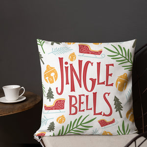 A pillow on a chair with a coffee mug on a table next to it. The white pillow features the words "Jingle Bells" in red in the center with Christmas illustrations surrounding the words. The Christmas illustrations are pine trees, ornaments, leaves and sleighs. The illustrations are in the colors yellow, light blue, red, and light and dark green. 