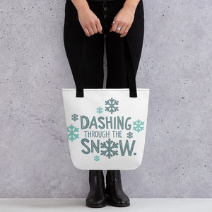 Someone holding a tote bag with black handles and a white fabric bag. The tote bag features the words "Dashing through the snow" in light and dark blue with snowflakes around the words. 