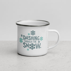 White enamel mug sitting on top of a tree branch with moss in the background. The design on the mug is featured in light and dark blue reading "Dashing through the snow" with the "O" of snow as a snowflake. There are also snowflakes surrounding the design. 