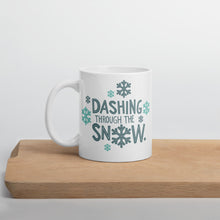 Load image into Gallery viewer, A white mug sitting on a light wood cutting board. The illustrated design says &quot;Dashing throug the snow&quot; with the &quot;O&quot; of snow as a snowflake. There are snowflakes surrounding the words. The words and snowflakes are in dark and light blue. 