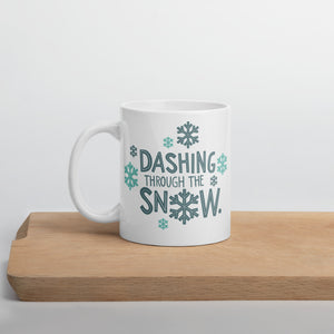 A white mug sitting on a light wood cutting board. The illustrated design says "Dashing throug the snow" with the "O" of snow as a snowflake. There are snowflakes surrounding the words. The words and snowflakes are in dark and light blue. 